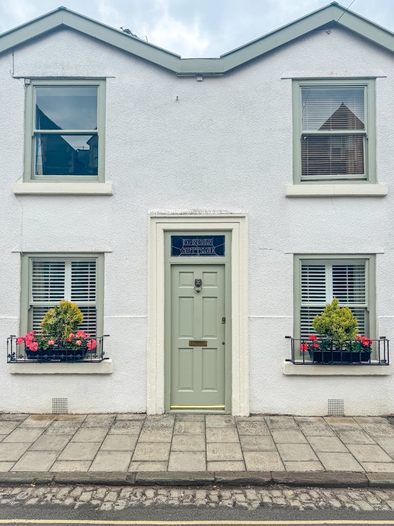 Charming white English cottage with sage door, green window trim, and vibrant flowers.